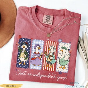 Pu&ograve; includere: T-shirt color cremisi con una grafica di quattro oche vestite con abiti patriottici. Le oche sono davanti a sfondi rossi, bianchi e blu. Il testo in corsivo "Just an independent goose" &egrave; sotto.