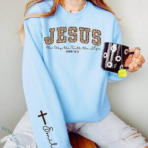 May include: Light blue sweatshirt with the word "JESUS" in leopard print lettering. The text "the Way, the Truth, the Life JOHN 14:6" is below the word "JESUS". A black cross and the name "Emily" are printed on the left sleeve.