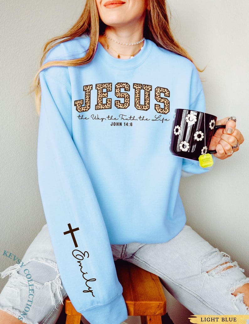 May include: Light blue sweatshirt with the word "JESUS" in leopard print lettering. The text "the Way, the Truth, the Life JOHN 14:6" is below the word "JESUS". A black cross and the name "Emily" are printed on the left sleeve.