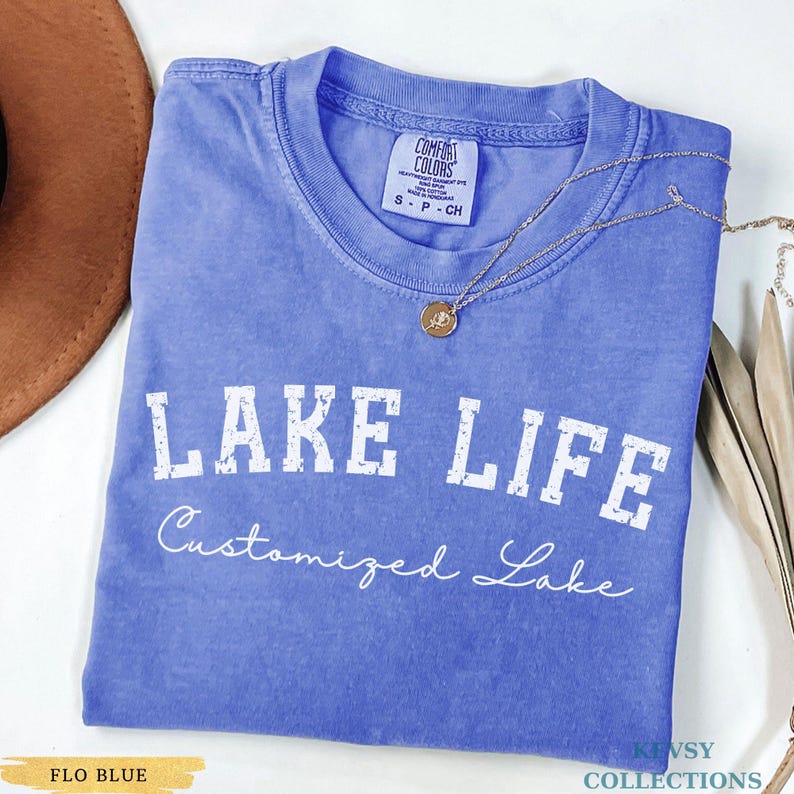 May include: A blue t-shirt with the words "LAKE LIFE" in white block letters and "Customized Lake" in cursive. A gold necklace with a pendant is on the shirt. The shirt is labeled "Flo Blue" and is a Comfort Colors brand.