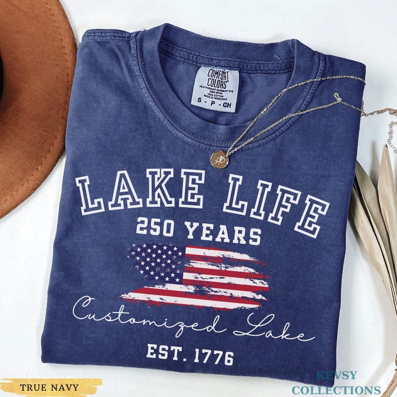 Puede incluir: Una camiseta azul marino con las palabras "LAKE LIFE" y "250 YEARS" en blanco. Debajo hay un dise&ntilde;o de la bandera estadounidense. Tambi&eacute;n se ve el texto "Customized Lake" y "EST. 1776". Un collar dorado est&aacute; sobre la camiseta.