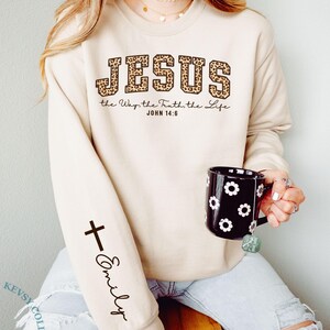 May include: Sand-colored sweatshirt with the word "JESUS" in leopard print lettering. The text "the Way, the Truth, the Life JOHN 14:6" is below. A black cross and the name "Emily" are on the sleeve. A black mug with white flower designs is held.