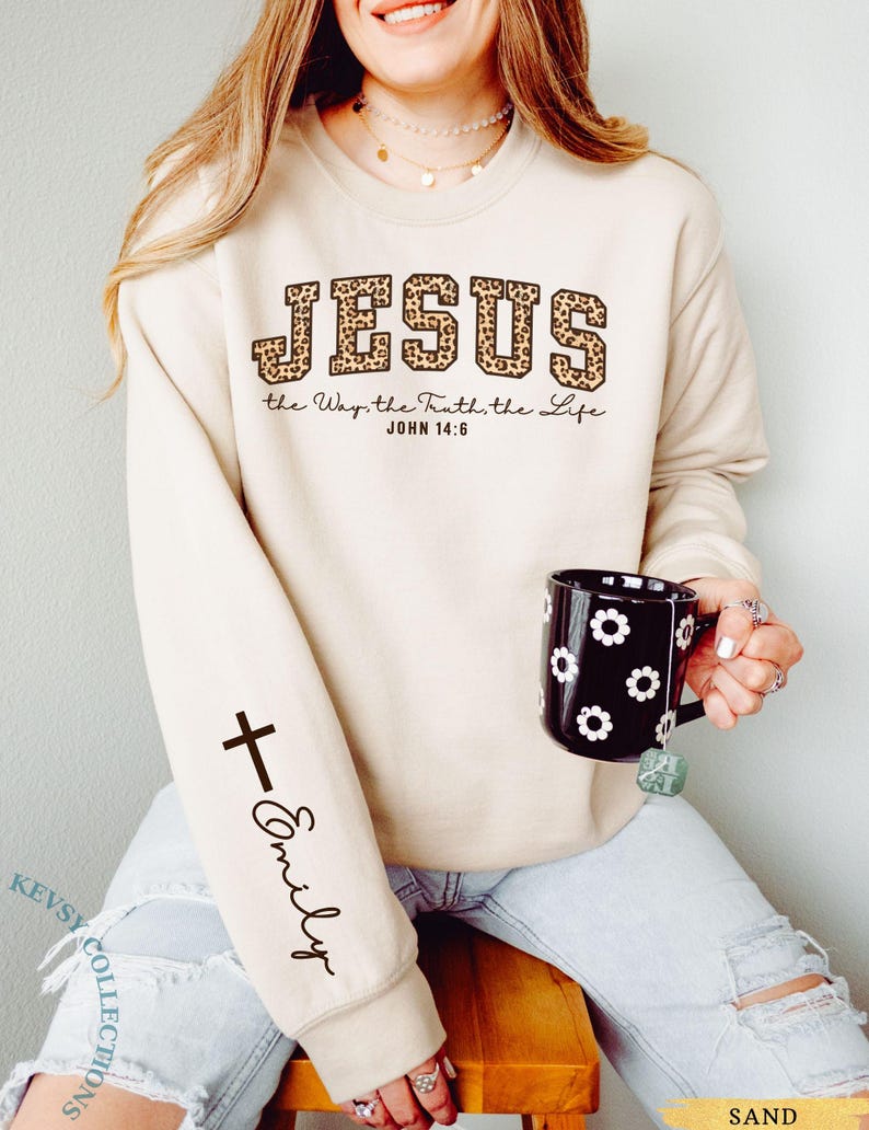 May include: Sand-colored sweatshirt with the word "JESUS" in leopard print lettering. The text "the Way, the Truth, the Life JOHN 14:6" is below. A black cross and the name "Emily" are on the sleeve. A black mug with white flower designs is held.