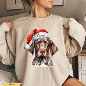May include: Sand-colored sweatshirt featuring a watercolor illustration of a dog wearing a Santa hat. The hat is red with white polka dots and a white pom-pom. The dog's name, "Luna," is printed on the hat. The sweatshirt is a cozy, casual style.