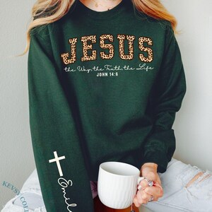 May include: Forest green sweatshirt with the word "JESUS" in leopard print, and the text "the Way, the Truth, the Life, John 14:6". A white cross and the name "Emily" are on the sleeve. A white mug is held in the hand.