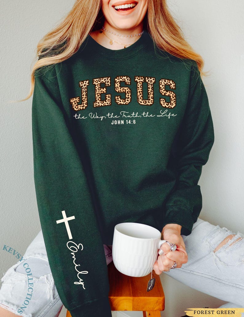 May include: Forest green sweatshirt with the word "JESUS" in leopard print, and the text "the Way, the Truth, the Life, John 14:6". A white cross and the name "Emily" are on the sleeve. A white mug is held in the hand.