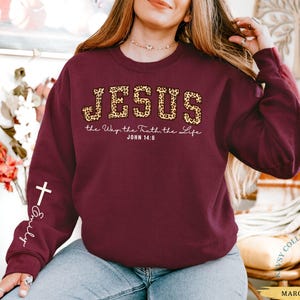 May include: Maroon sweatshirt with the word "JESUS" in leopard print, along with the text "the Way, the Truth, the Life, John 14:6." A white cross and the name "Emily" are on the left sleeve. The word "MAROON" is at the bottom.