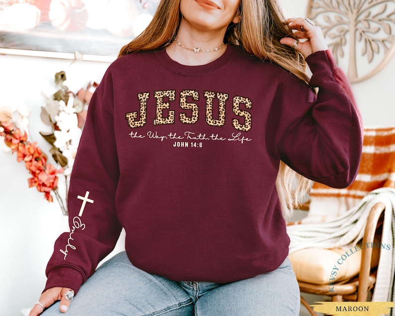 May include: Maroon sweatshirt with the word "JESUS" in leopard print, along with the text "the Way, the Truth, the Life, John 14:6." A white cross and the name "Emily" are on the left sleeve. The word "MAROON" is at the bottom.