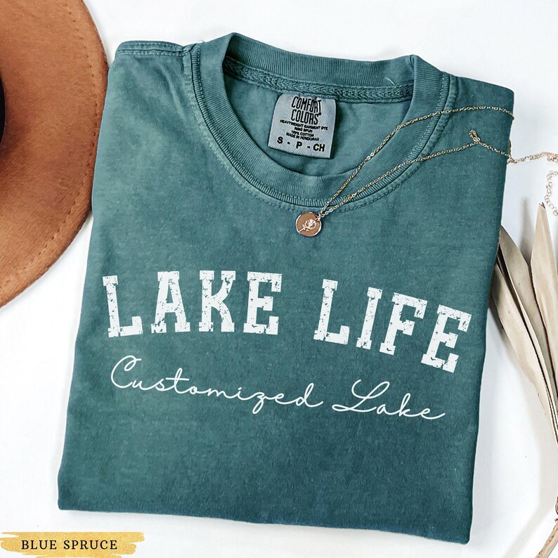 May include: A blue spruce t-shirt with the words "LAKE LIFE" in white block letters. Below the text is the cursive text "Customized Lake". The shirt has a crew neck and a small tag on the upper back. A gold necklace with a pendant is draped across the shirt.