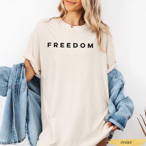 May include: Ivory-colored t-shirt with the word "FREEDOM" printed in black. The shirt is worn with a light blue denim jacket. The model is wearing a gold necklace.