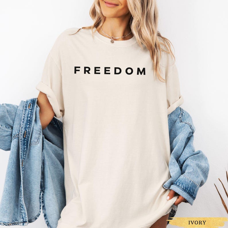 May include: Ivory-colored t-shirt with the word "FREEDOM" printed in black. The shirt is worn with a light blue denim jacket. The model is wearing a gold necklace.