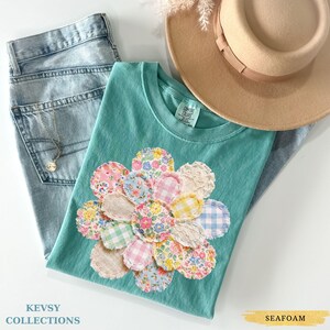May include: A teal t-shirt with a floral patchwork design, alongside light wash jeans and a tan hat. The patchwork flower is made of various patterned fabrics. The text "KEVSY COLLECTIONS" is visible.