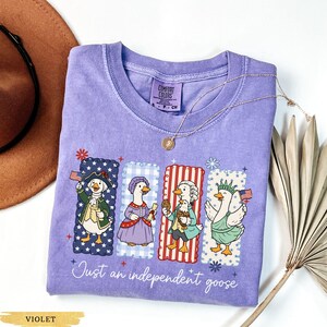Pu&ograve; includere: Maglietta Comfort Colors viola con quattro oche dei cartoni animati vestite con abiti patriottici. Ogni oca &egrave; in una cornice rettangolare con un motivo diverso. Il testo "Just an independent goose" &egrave; scritto in corsivo sotto.