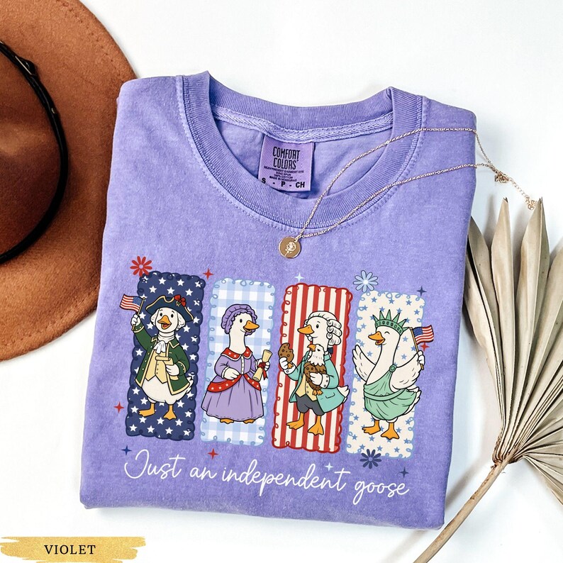 Pu&ograve; includere: Maglietta Comfort Colors viola con quattro oche dei cartoni animati vestite con abiti patriottici. Ogni oca &egrave; in una cornice rettangolare con un motivo diverso. Il testo "Just an independent goose" &egrave; scritto in corsivo sotto.