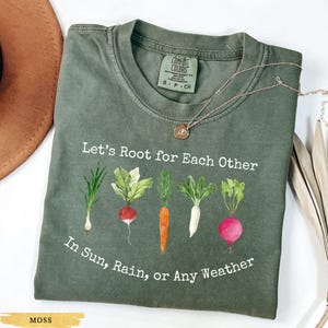 Könnte beinhalten: Moosgrünes T-Shirt mit dem Text "Let's Root for Each Other" und "In Sun, Rain, or Any Weather." Das Shirt zeigt eine Aquarellillustration verschiedener Wurzelgemüse. Eine goldene Halskette und ein brauner Hut sind ebenfalls zu sehen.