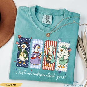 Pu&ograve; includere: T-shirt verde acqua con un design di oca patriottica. La maglietta presenta quattro oche dei cartoni animati vestite con abiti storici, ognuna con una bandiera americana o una pergamena. Il testo "Just an independent goose" &egrave; stampato sotto.