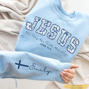 Peut inclure: Sweat-shirt bleu clair avec le mot "JESUS" en grandes lettres à motifs floraux. Le texte "the Way, the Truth, the Life, John 14:6" est en dessous. La manche présente une croix et le nom "Emily".