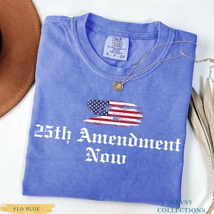 May include: A blue t-shirt with the text "25th Amendment Now" in white gothic font. The shirt features a distressed American flag graphic. A gold necklace with a pendant is draped on the shirt. The shirt is labeled "Flo Blue".