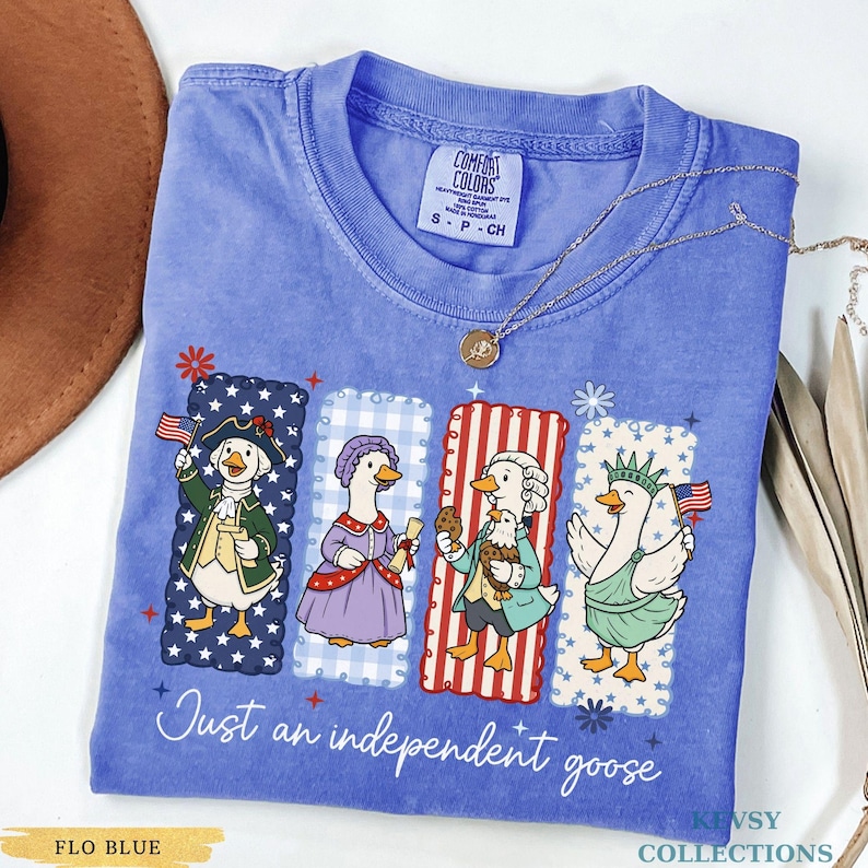 Pu&ograve; includere: T-shirt Comfort Colors blu Flo con una grafica di quattro oche vestite con abiti patriottici. Il testo "Just an independent goose" &egrave; stampato sotto la grafica. Una collana d'oro &egrave; drappeggiata sulla maglietta.