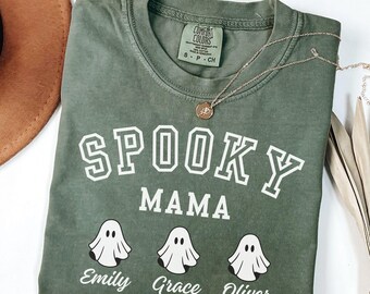 May include: A moss green t-shirt featuring the text "SPOOKY MAMA" in white. Below are three ghost illustrations and the names "Emily", "Grace", and "Oliver". A gold necklace and a brown hat are also visible.