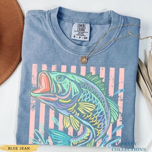 Op de afbeelding: Een blauw spijkerkleurig t-shirt met een afbeelding van een vis in tinten groen, geel en paars. Het ontwerp bevat verticale roze strepen en waterspatten. Het shirt heeft een Comfort Colors label en er ligt een gouden ketting op.