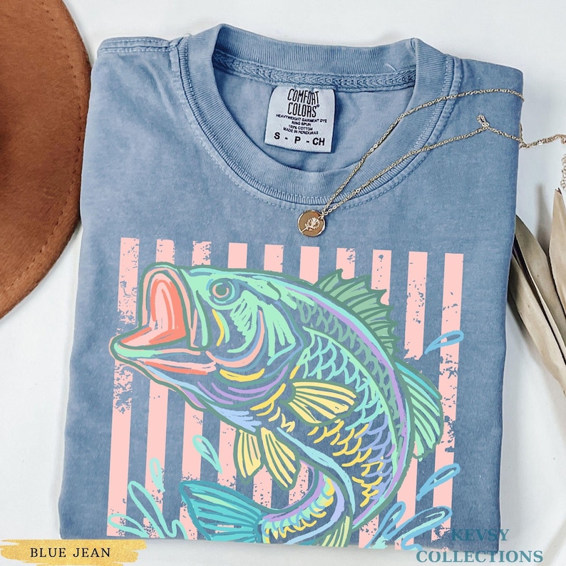 Op de afbeelding: Een blauw spijkerkleurig t-shirt met een afbeelding van een vis in tinten groen, geel en paars. Het ontwerp bevat verticale roze strepen en waterspatten. Het shirt heeft een Comfort Colors label en er ligt een gouden ketting op.