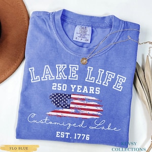 Puede incluir: Una camiseta azul con las palabras "LAKE LIFE" y "250 YEARS" en letras blancas. Debajo, una bandera estadounidense estilizada y las palabras "Customized Lake" y "EST. 1776". Un collar dorado est&aacute; drapeado sobre la camiseta.