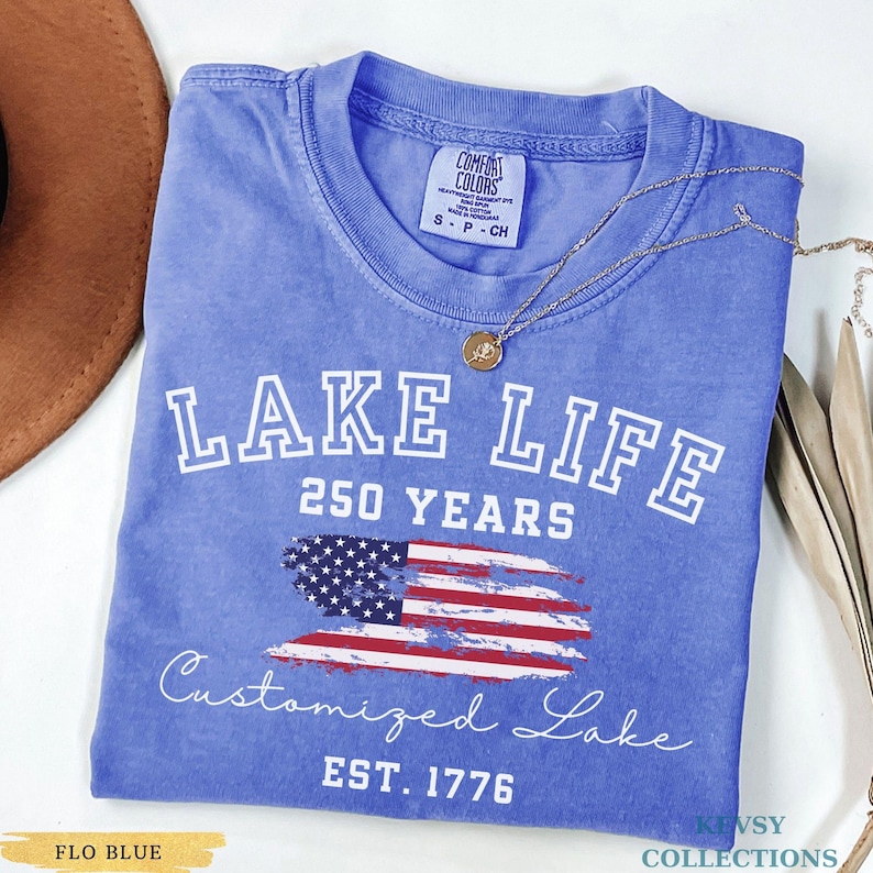 Puede incluir: Una camiseta azul con las palabras "LAKE LIFE" y "250 YEARS" en letras blancas. Debajo, una bandera estadounidense estilizada y las palabras "Customized Lake" y "EST. 1776". Un collar dorado est&aacute; drapeado sobre la camiseta.