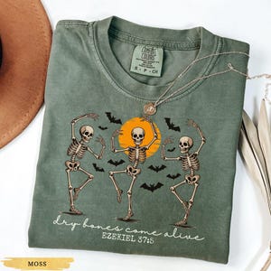 May include: Moss green t-shirt featuring a graphic of three dancing skeletons in front of an orange sun with black bats. The text "dry bones come alive" and "Ezekiel 37:5" are printed below the graphic. A silver necklace is draped over the shirt.