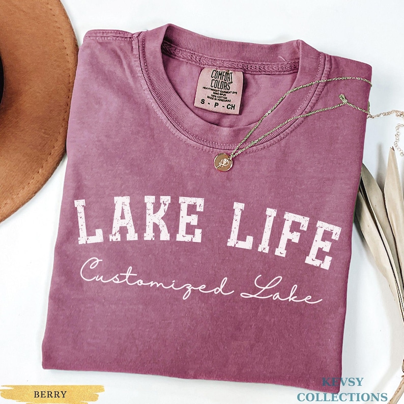 May include: A berry-colored t-shirt with the words "LAKE LIFE" in white, distressed lettering. Below, the text "Customized Lake" is written in a cursive font. A gold necklace with a pendant rests on the shirt. The shirt has a Comfort Colors tag.