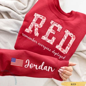 Könnte beinhalten: Ein rotes Sweatshirt mit Rundhalsausschnitt und dem Wort RED in großen Blockbuchstaben, die mit einem Blumenmuster gefüllt sind. Darunter steht der Satz remember everyone deployed. Der Ärmel zeigt eine US-Flagge und den Namen Jordan in Schreibschrift.