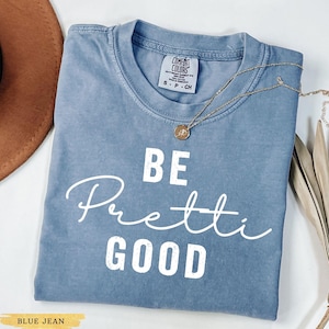 May include: A blue jean colored t-shirt with the words "BE Pretti GOOD" in white script. The shirt has a crew neck and short sleeves. A gold necklace with a pendant is draped across the shirt.