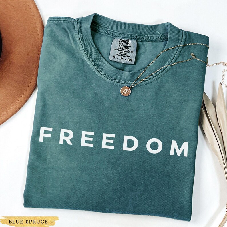 May include: A blue spruce t-shirt with the word "FREEDOM" in white block letters. The shirt has a crew neck and short sleeves. A gold necklace with a pendant is draped across the shirt.
