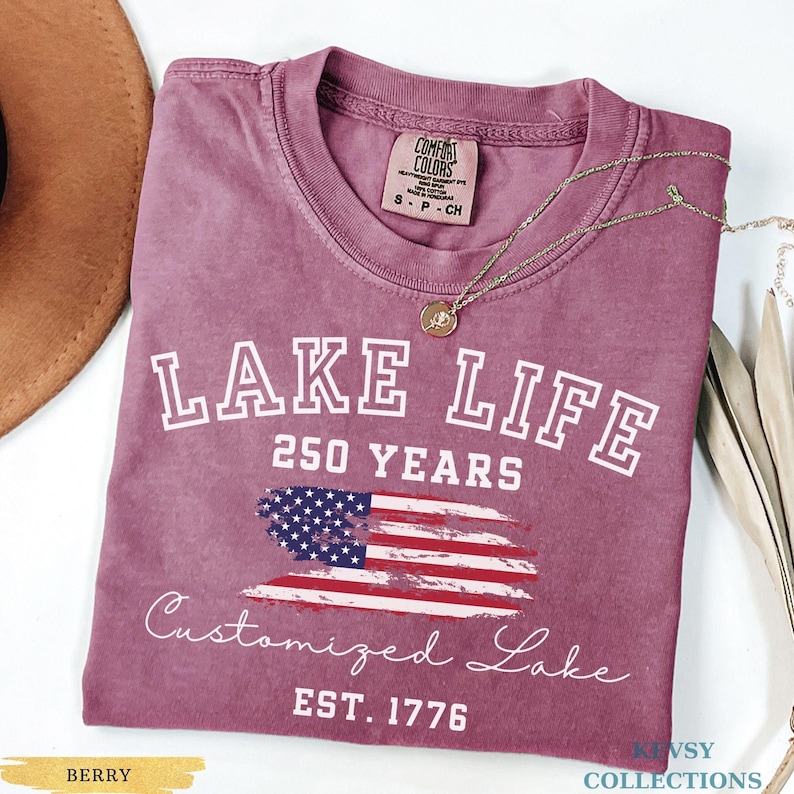 Puede incluir: Camiseta de color bayas con las palabras "LAKE LIFE" y "250 YEARS" en letras blancas. Debajo hay un dise&ntilde;o de la bandera estadounidense. Tambi&eacute;n se ve el texto "Customized Lake" y "EST. 1776". Un collar dorado est&aacute; sobre la camiseta.