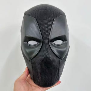 Puede incluir: Una máscara negra de Deadpool con cubiertas oculares de malla blanca. La máscara tiene una superficie texturizada y está diseñada para cubrir toda la cara. Las formas de los ojos están delineadas en un material liso y mate.