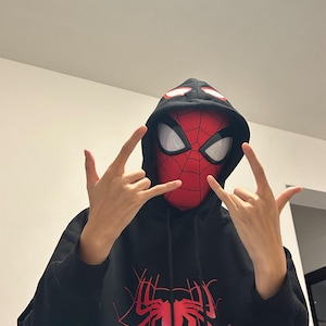 Op de afbeelding: Een zwarte hoodie met een rood Spider-Man-masker. Het masker is rood met een zwart webpatroon en witte ooglenzen. De hoodie heeft een rode spinnenafbeelding op de voorkant.