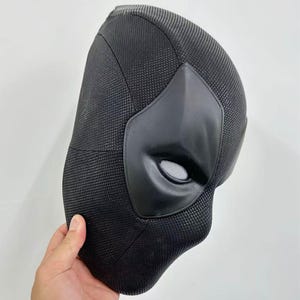 Puede incluir: Una m&aacute;scara negra de Deadpool con un acabado de tela texturizada. La m&aacute;scara presenta un &aacute;rea de ojos y boca negra mate y lisa con una cubierta ocular de malla. La m&aacute;scara est&aacute; sujeta por una mano.