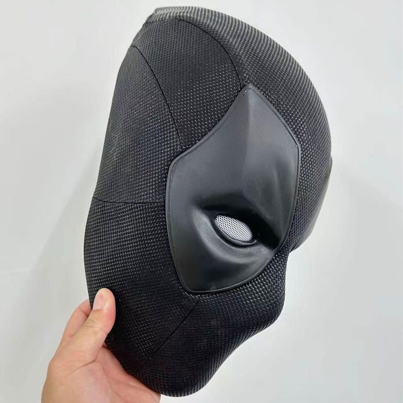 Puede incluir: Una m&aacute;scara negra de Deadpool con un acabado de tela texturizada. La m&aacute;scara presenta un &aacute;rea de ojos y boca negra mate y lisa con una cubierta ocular de malla. La m&aacute;scara est&aacute; sujeta por una mano.