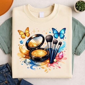 Puede incluir: Sudadera de color crema con un diseño de maquillaje. El diseño incluye una polvera con espejo, brochas de maquillaje, mariposas y gemas. El diseño es en tonos dorados, azules, rosas y naranjas.