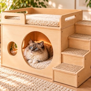 Può includere: Una cuccia per gatti in legno chiaro con un grande ingresso circolare, una finestra laterale rotonda più piccola e una piattaforma superiore con un cuscino beige testurizzato. Tre gradini integrati con moquette beige portano in cima.