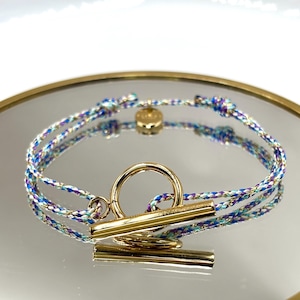 Peut inclure: Un bracelet tressé avec un fermoir à bascule doré. Le bracelet est composé d'un cordon multicolore avec des fils bleus, violets et dorés. Le fermoir est une barre et un anneau dorés. Le bracelet est présenté sur une surface réfléchissante.
