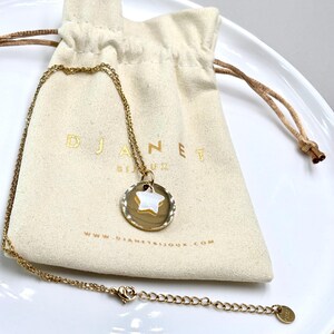Puede incluir: Un collar dorado con un colgante de estrella y una bolsa con cordón a juego. El colgante presenta una estrella blanca dentro de un marco circular. La bolsa tiene el nombre de la marca "DJANET" y la dirección del sitio web.