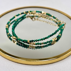 Peut inclure: Un collier à plusieurs rangs composé de petites perles vert foncé facettées et de perles géométriques dorées. Le collier est présenté sur une surface réfléchissante avec un bord doré, mettant en valeur son design délicat et son contraste de couleurs.