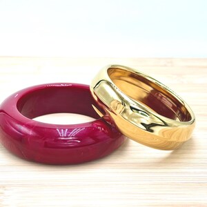 Può includere: Una coppia di bracciali rigidi spessi composta da un bracciale in metallo color oro lucido e un bracciale in resina color magenta intenso con una texture marmorizzata. Il bracciale color oro presenta una superficie liscia e riflettente, mentre quello magenta è opaco.