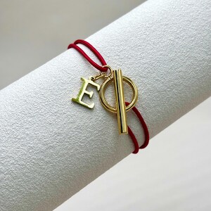Può includere: Un braccialetto con cordino rosso con chiusura color oro e un ciondolo a forma di lettera "E" dorata. Il braccialetto è presentato su una superficie bianca strutturata. Il design del braccialetto è semplice ed elegante.