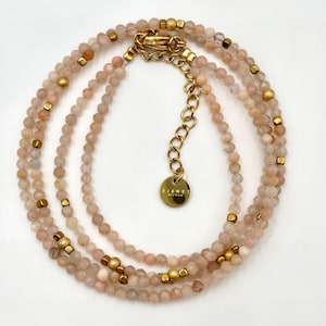Può includere: Una delicata collana con più fili di piccole perline rosa chiaro. Accenti color oro e una chiusura aggiungono contrasto. Un piccolo ciondolo rotondo dorato con la scritta "DJANET BIJOUX" pende vicino alla chiusura.