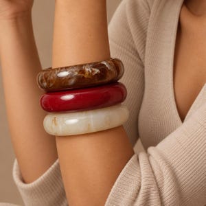 Può includere: Un set di tre bracciali rigidi spessi e arrotondati impilati insieme. Il bracciale superiore è marrone marmorizzato, quello centrale è rosso intenso a tinta unita e quello inferiore è bianco sporco marmorizzato. Ogni bracciale ha una finitura liscia.