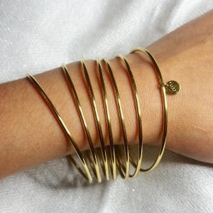 Peut inclure: Plusieurs bracelets dorés fins et ronds sur un poignet. Ils présentent une finition lisse et brillante. L'un des bracelets est orné d'un petit charme rond. L'ensemble est présenté sur un fond blanc.