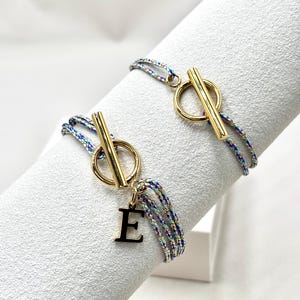 Könnte beinhalten: Zwei Armbänder mit goldfarbenen Knebelverschlüssen und mehrfarbigen Kordeln. Ein Armband hat einen schwarzen Buchstaben "E" Anhänger. Die Armbänder werden auf einer weißen, strukturierten Oberfläche präsentiert.
