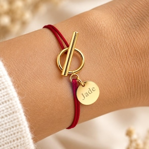 Può includere: Un bracciale in cordino rosso con chiusura a T dorata e un piccolo ciondolo rotondo dorato inciso con il nome Jade. Il bracciale è fissato con una chiusura a barra e anello, caratterizzato da una finitura metallica lucida.
